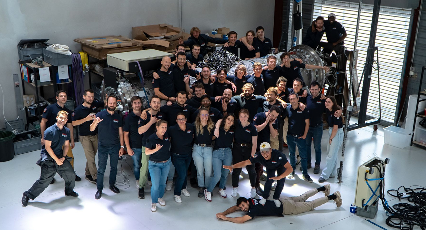 Large group photo of the HyPrSpace team in their workshop, smiling, gesturing playfully, and posing in front of propulsion equipment. A strong expression of company culture.