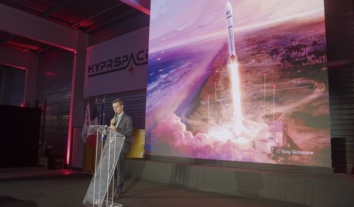 A speaker addresses guests at the opening of HyPrSpace’s new factory, beside an illustration of Baguette One.